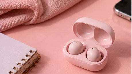 Pastel pink wireless earbuds in an open charging case on a pink desk beside a notebook.