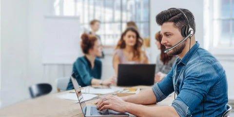 Man wearing Wantek wired headset working on laptop in modern office with colleagues in background