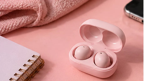 Pastel pink wireless earbuds in an open charging case on a pink desk beside a notebook.